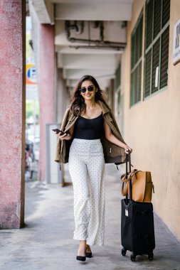 young and attractive Asian Indian woman books a ride through her ride hailing app on her smartphone. She is standing in a walkway with her luggage and is stylishly dressed in work wear and shades clipart