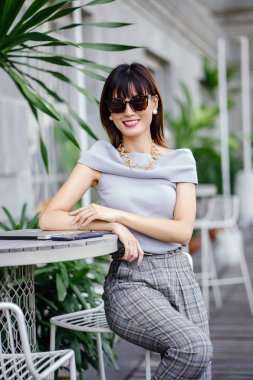 Açık havada şık bir yerde oturan bir emin, şık ve çekici orta yaşlı Çinli Asyalı kadın portresi. O bir pohpohlama gri kıyafeti giyiyor.