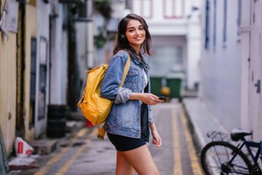 Bir genç ve çekici Hint Asyalı kadın bir kot ceket ve sarı sırt çantası onun kulaklıklar üzerinde müzik keyfi portresi