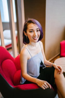 Profesyonel elbiseli ve boyalı mor saçlı genç, ince ve zarif bir Asyalı kadın çalışma odasında kırmızı bir koltukta oturuyor. İçtenlikle gülümsüyor..