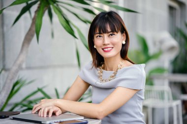 Açık havada şık bir yerde oturan bir emin, şık ve çekici orta yaşlı Çinli Asyalı kadın portresi. O bir pohpohlama gri kıyafeti giyiyor.