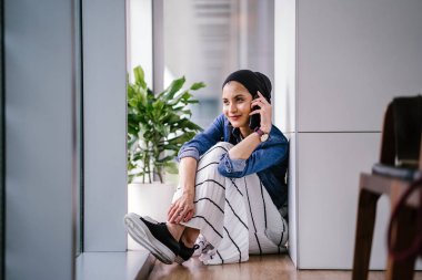 Genç bir Ortadoğulu Müslüman kadın gün boyunca bir ofiste, ortak çalışma alanında veya kütüphanede oturuyor ve akıllı telefonunu kullanıyor. Başörtüsü takıyor (başörtüsü)).