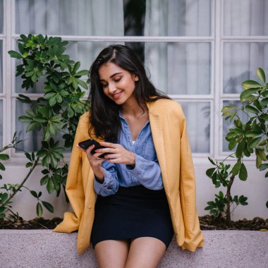 güzel, genç ve şık Hint Asyalı kadın manifatura onun Smartphone olarak o gün karşı bir çıkıntıya doğru eğilir. O rahat kıyafetler üzerinde bir retro sarı ceket giyiyor