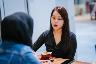 Çinli bir Asyalı kadın ve Müslüman bir kadın kafede eğleniyorlar.