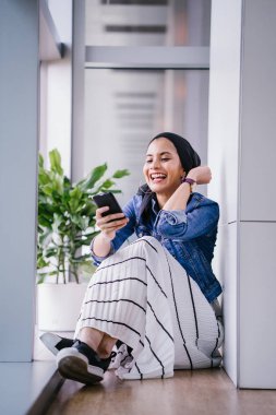Genç bir Ortadoğulu Müslüman kadın gün boyunca bir ofiste, ortak çalışma alanında veya kütüphanede oturuyor ve akıllı telefonunu kullanıyor. Başörtüsü takıyor (başörtüsü)).