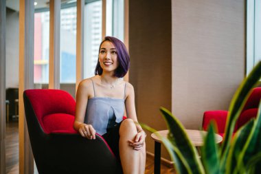 Profesyonel elbiseli ve boyalı mor saçlı genç, ince ve zarif bir Asyalı kadın çalışma odasında kırmızı bir koltukta oturuyor. İçtenlikle gülümsüyor..