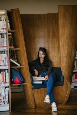 Genç, güzel ve çekici Çinli Asyalı değişim öğrencisi kütüphanedeki kahverengi bir kulübede okuyor ve çalışıyor..