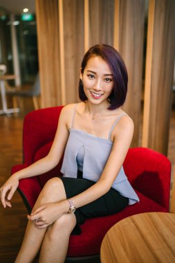 Profesyonel elbiseli ve boyalı mor saçlı genç, ince ve zarif bir Asyalı kadın çalışma odasında kırmızı bir koltukta oturuyor. İçtenlikle gülümsüyor..