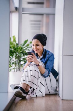 Genç bir Ortadoğulu Müslüman kadın gün boyunca bir ofiste, ortak çalışma alanında veya kütüphanede oturuyor ve akıllı telefonunu kullanıyor. Başörtüsü takıyor (başörtüsü)).