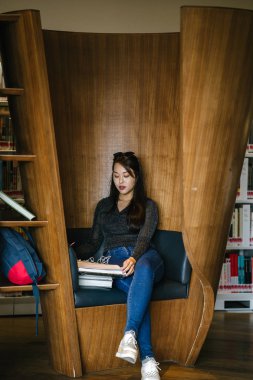 Genç, güzel ve çekici Çinli Asyalı değişim öğrencisi kütüphanedeki kahverengi bir kulübede okuyor ve çalışıyor..