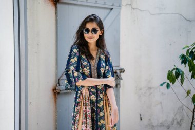 uzun boylu, zarif ve güzel Hint Asyalı kadın yaz elbise ve güneş gözlüğü gülümsüyor o pozlar gün boyunca bir arka sokakta bir sokakta olarak içinde