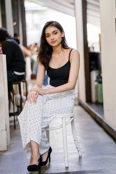 Bir uzun boylu, zarif ve güzel Hint gün boyunca şehrin bir kafede bir sandalyede oturan kız en iyi ve uzun beyaz siyah pantolon Asya portresi