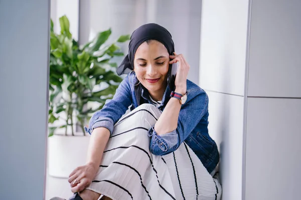 一名年轻的中东穆斯林妇女白天坐在办公室 工作空间或图书馆的角落里 用她的智能手机 她戴着头巾 — 图库照片