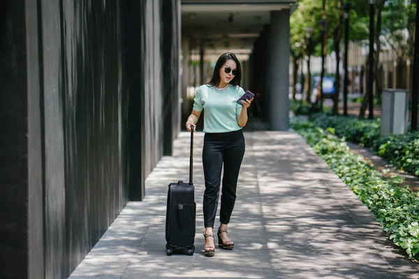 Portrait of an elegant and beautiful young Asian woman waiting on the side of the street for her ride that she booked via a ride-hailing app. She is holding onto a luggage suitcase on wheels.