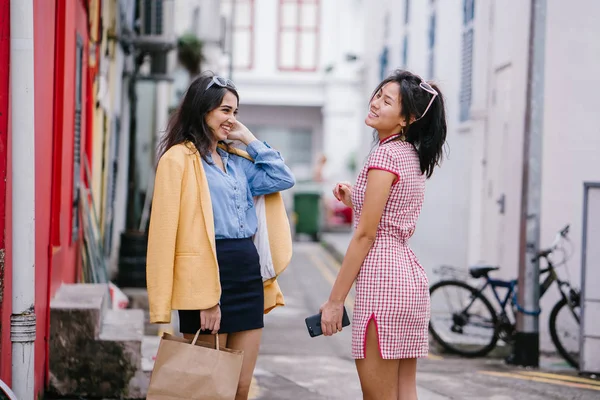 two close friends walking together down an alley. They are both wearing trendy retro clothing and holding hands as they walk happily together. One is Chinese, the other Indian