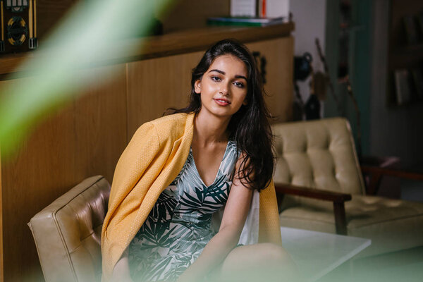  fashionable, beautiful and young Indian Asian woman in a dress and a yellow jacket draped over her shoulders. She is sitting on a chair in a stylish interior