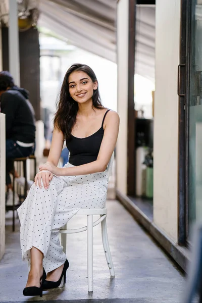 Bir uzun boylu, zarif ve güzel Hint gün boyunca şehrin bir kafede bir sandalyede oturan kız en iyi ve uzun beyaz siyah pantolon Asya portresi