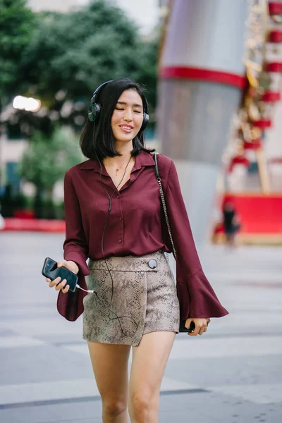 beautiful Korean woman in an elegant blouse and shorts dancing as she walks down a street in the city with her headphones on