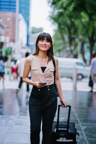 attractive Asian Chinese woman books a ride through her ride hailing app on her smartphone. She is walking with her luggage and is stylishly dressed in work wear