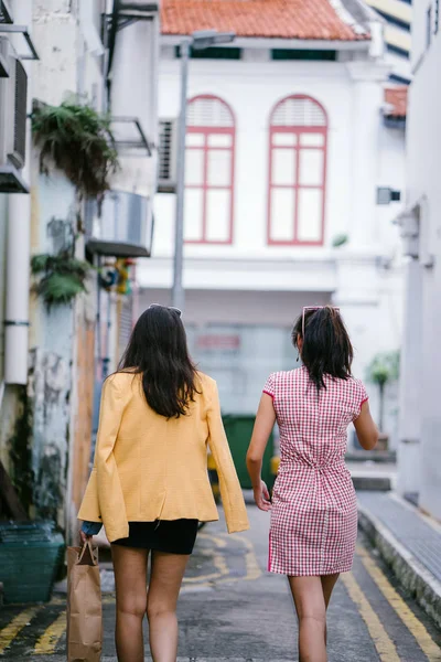 two close friends walking together down an alley. They are both wearing trendy retro clothing and holding hands as they walk happily together. One is Chinese, the other Indian