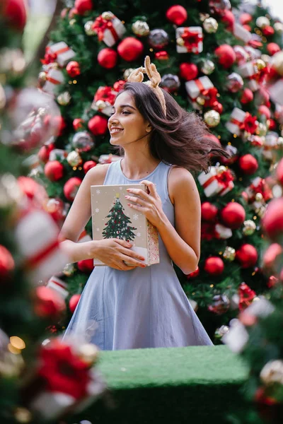 Genç, güzel ve mutlu bir Hint Asyalı kadın şenlikli Noel portresi bir hediye bir Noel ağacı önünde tutan gülümseyerek.