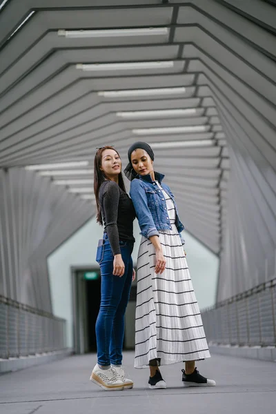 Two friends of different ethnicity on a bridge during the day. One is a Malay Muslim woman and the other is Chinese Asian. They are both young, energetic and attractive.
