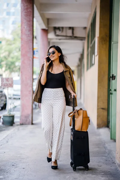 young and attractive Asian Indian woman books a ride through her ride hailing app on her smartphone. She is standing in a walkway with her luggage and is stylishly dressed in work wear and shades