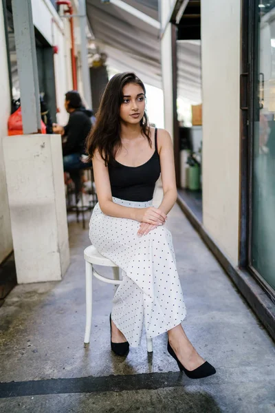 Bir uzun boylu, zarif ve güzel Hint gün boyunca şehrin bir kafede bir sandalyede oturan kız en iyi ve uzun beyaz siyah pantolon Asya portresi