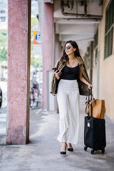 young and attractive Asian Indian woman books a ride through her ride hailing app on her smartphone. She is standing in a walkway with her luggage and is stylishly dressed in work wear and shades