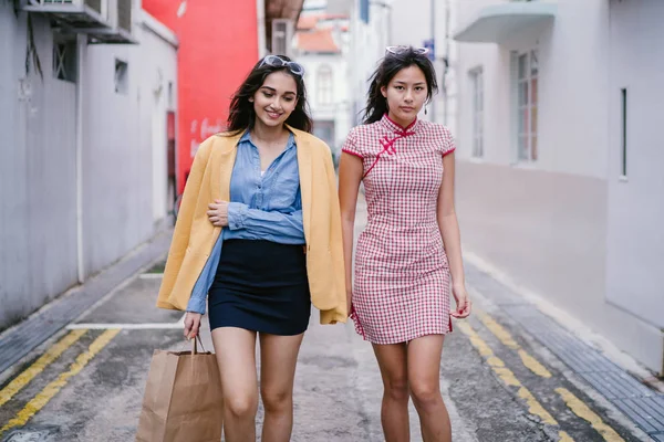 two close friends walking together down an alley. They are both wearing trendy retro clothing and holding hands as they walk happily together. One is Chinese, the other Indian