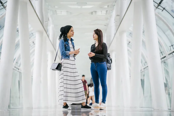 Two friends of different ethnicity stand next to one another on a bridge during the day. One is a Malay Muslim woman and the other is Chinese Asian. They are both young, energetic and attractive.