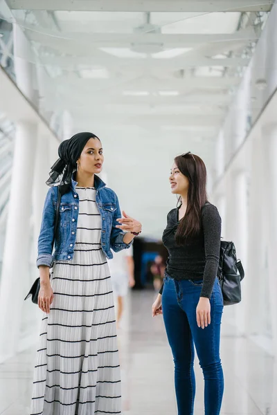 Two friends of different ethnicity stand next to one another on a bridge during the day. One is a Malay Muslim woman and the other is Chinese Asian. They are both young, energetic and attractive.