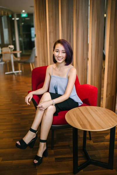 Profesyonel elbiseli ve boyalı mor saçlı genç, ince ve zarif bir Asyalı kadın çalışma odasında kırmızı bir koltukta oturuyor. İçtenlikle gülümsüyor..