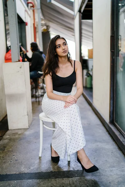 Bir uzun boylu, zarif ve güzel Hint gün boyunca şehrin bir kafede bir sandalyede oturan kız en iyi ve uzun beyaz siyah pantolon Asya portresi