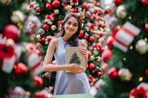 Genç, güzel ve mutlu bir Hint Asyalı kadın şenlikli Noel portresi bir hediye bir Noel ağacı önünde tutan gülümseyerek.
