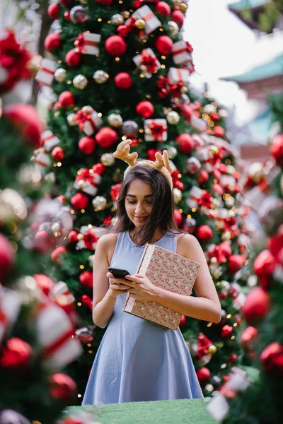 Genç, güzel ve mutlu bir Hint Asyalı kadın şenlikli Noel portresi bir hediye bir Noel ağacı önünde tutan gülümseyerek.