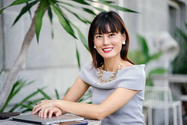 Açık havada şık bir yerde oturan bir emin, şık ve çekici orta yaşlı Çinli Asyalı kadın portresi. O bir pohpohlama gri kıyafeti giyiyor.