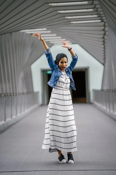 Güzel bir köprünün (geometrik desenler) geçidinde duran uzun, zarif ve ince bir Orta Doğulu kadının tam vücut portresi. Kollarını açmış ve komik bir yüz ifadesi takınıyor..