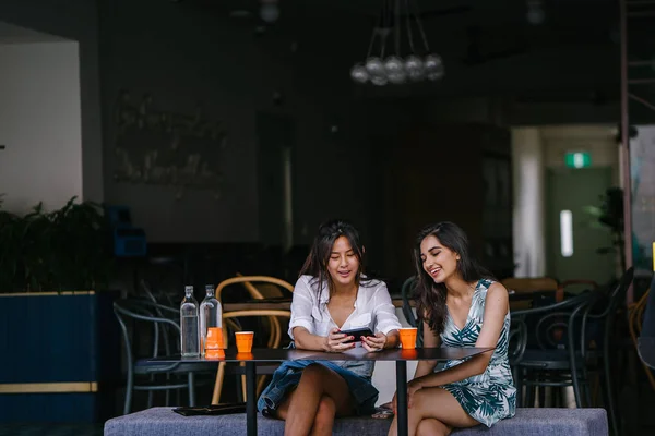 Two close friends (women) sit in a cafe during the day and watch a video together on a smartphone. They are both smiling as they enjoy the video and each other\'s company.