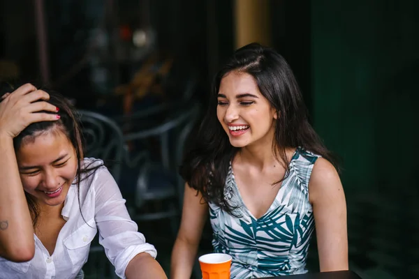 Two close friends (women) sit in a cafe during the day and watch a video together on a smartphone. They are both smiling as they enjoy the video and each other\'s company.