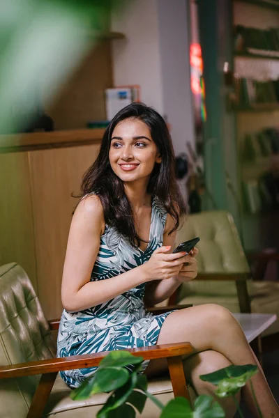 young and beautiful Indian Asian woman sits on a chair in a well appointed interior. She is checking her smartphone as she enjoys the day.