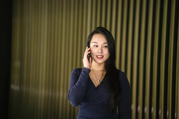 一个年轻漂亮的亚洲中国妇女的肖像 穿着优雅优雅的蓝色连衣裙 与现代金色特色墙 她穿着很酷的太阳镜 在她的智能手机上说话 — 图库照片