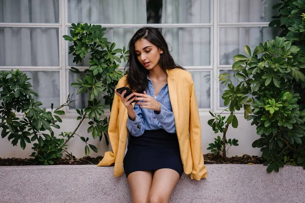 güzel, genç ve şık Hint Asyalı kadın manifatura onun Smartphone olarak o gün karşı bir çıkıntıya doğru eğilir. O rahat kıyafetler üzerinde bir retro sarı ceket giyiyor