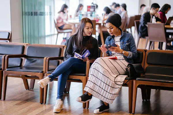 两个年轻的女性朋友坐在长椅上看书和讨论 一个是马来族穆斯林妇女 另一个是中国妇女 — 图库照片