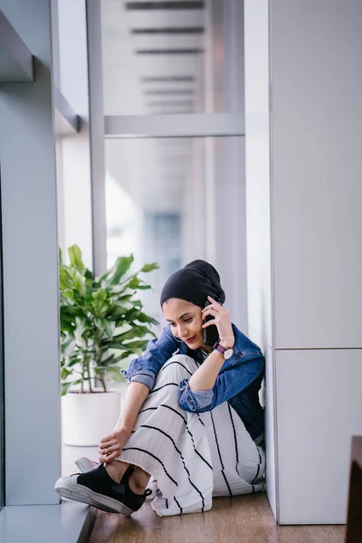 A young Middle-Eastern Muslim woman sits in a corner of an office, coworking space or library during the day and is using her smartphone. She is wearing a head scarf (hijab).
