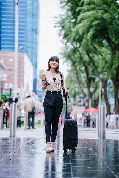 attractive Asian Chinese woman books a ride through her ride hailing app on her smartphone. She is walking with her luggage and is stylishly dressed in work wear