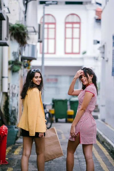 two close friends walking together down an alley. They are both wearing trendy retro clothing and holding hands as they walk happily together. One is Chinese, the other Indian