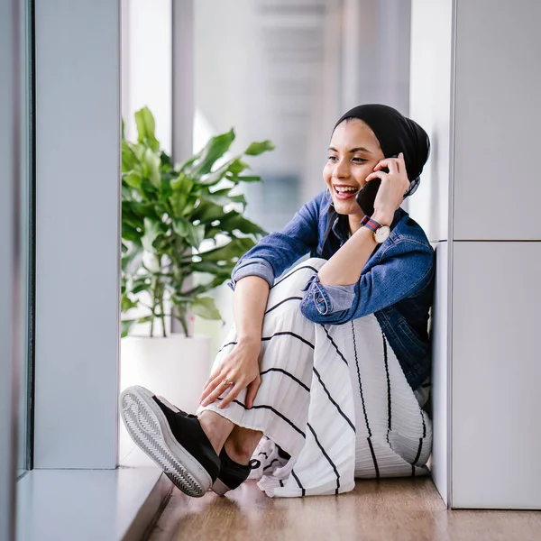 A young Middle-Eastern Muslim woman sits in a corner of an office, coworking space or library during the day and is using her smartphone. She is wearing a head scarf (hijab).