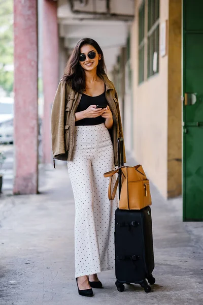 young and attractive Asian Indian woman books a ride through her ride hailing app on her smartphone. She is standing in a walkway with her luggage and is stylishly dressed in work wear and shades