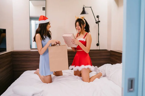 Two young Asian girls (one Indian, one Chinese) exchange Christmas gifts. One is wearing a red Santarina outfit and the other a blue dress with a Santa hat. They are both smiling happily.